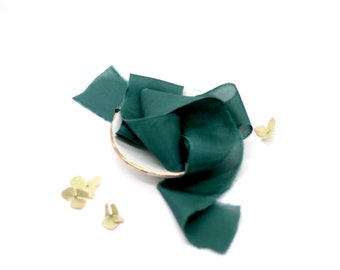 May include: A teal blue silk ribbon is arranged on a white plate with a gold rim. The ribbon is folded and twisted, creating a decorative display. There are three small green leaves scattered around the plate.