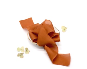 May include: A rust-colored silk ribbon is arranged in a loose bow on a white surface. The ribbon is soft and flowing, with a subtle sheen.