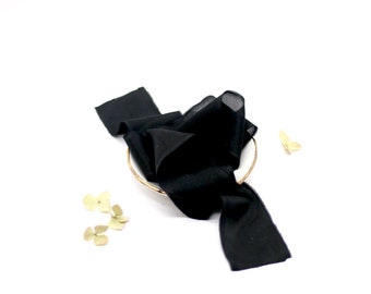May include: A black silk ribbon is draped over a gold-colored ring, with a few dried, light-colored flowers and a small gold-colored butterfly nearby. The ribbon is bunched up, creating soft folds and shadows against the white background.