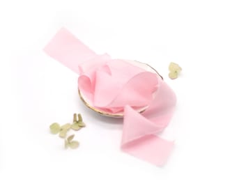 May include: A pale pink silk ribbon is coiled on a small white plate with a gold rim. The ribbon is soft and delicate, and it would be perfect for adding a touch of elegance to any project.