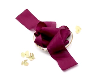 May include: A deep burgundy silk ribbon, folded and arranged in a small white bowl with a gold rim. The ribbon is soft and flowing, with a subtle sheen.