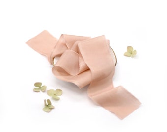 May include: A blush-colored silk ribbon is artfully arranged on a white surface, with delicate, dried green flowers scattered around. The ribbon's edges are slightly frayed, adding a touch of rustic charm to this elegant craft supply.