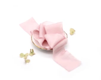 May include: A pale pink silk ribbon is draped over a small, white dish with a gold rim. Dried, light green flower buds are scattered around the dish on a white background. The ribbon has a soft, flowing texture.
