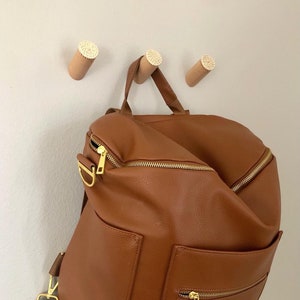 May include: A brown leather backpack with gold zippers and a front pocket. The backpack is hanging on three wooden hooks with white accents.