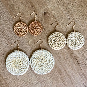 May include: Three pairs of woven rattan earrings. The earrings are round and have a natural, light brown colour. The earrings are on a wooden surface.