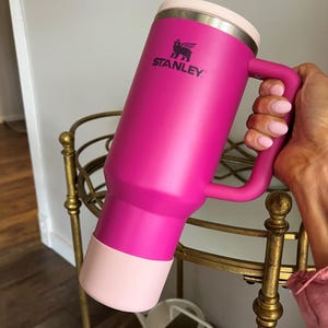 May include: A pink Stanley travel mug with a black logo and a pink lid with a straw. The mug has a handle and a pink silicone base.