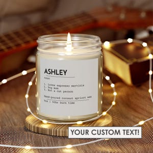 Puede incluir: Una vela blanca en un tarro de vidrio transparente con una etiqueta negra que dice "ASHLEY noun 1. Loves espresso martinis 2. Dog mom 3. Not a cat person Hand-poured coconut apricot wax 9oz | 50hr burn time". La vela está encendida y la llama es visible. La vela está sentada sobre una superficie de madera con una cadena de luces blancas envuelta alrededor.