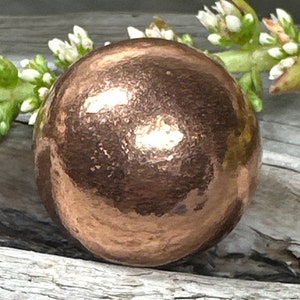 May include: A polished, spherical copper bead with a hammered texture. The surface of the round metal bead reflects light, creating bright highlights across its warm, metallic brown finish. The bead is positioned in front of small green buds.