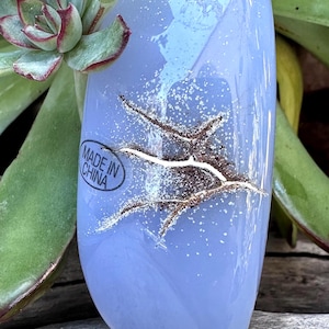 May include: A light blue, oval-shaped stone with a textured, crack-like design. The stone has a glossy finish and is surrounded by green succulent plants. The text "MADE IN CHINA" is visible on the stone.