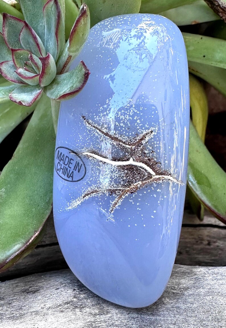 May include: A light blue, oval-shaped stone with a textured, crack-like design. The stone has a glossy finish and is surrounded by green succulent plants. The text "MADE IN CHINA" is visible on the stone.