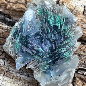 May include: A polished agate slice featuring a translucent, pale gray base with intricate, fan-shaped inclusions of vibrant green and dark metallic gray mineral formations radiating from the center, creating a natural, complex, and layered pattern.