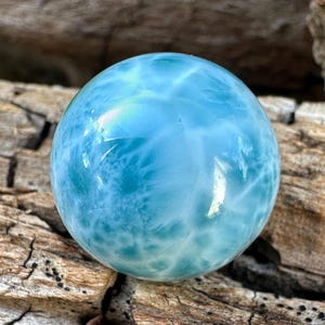 Può includere: Una pietra preziosa di Larimar lucida e sferica caratterizzata da una superficie liscia e brillante di un vibrante colore azzurro cielo con intricati motivi bianchi simili a nuvole, che mostrano variazioni naturali nella sua trama opaca.