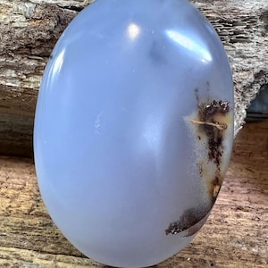 May include: A smooth, oval-shaped blue chalcedony gemstone with a polished, translucent surface. The stone features a distinct, dark brown, dendritic-like inclusion on the right side, contrasting with the soft, cool-toned blue body of the gem.