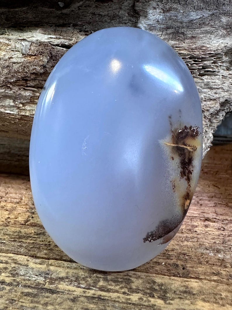May include: A smooth, oval-shaped blue chalcedony gemstone with a polished, translucent surface. The stone features a distinct, dark brown, dendritic-like inclusion on the right side, contrasting with the soft, cool-toned blue body of the gem.