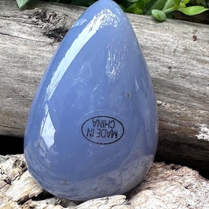 May include: A smooth, teardrop-shaped, pale blue stone. The stone has a glossy finish and a sticker that reads "MADE IN CHINA" in a black circular frame. The stone is resting on a piece of weathered wood.