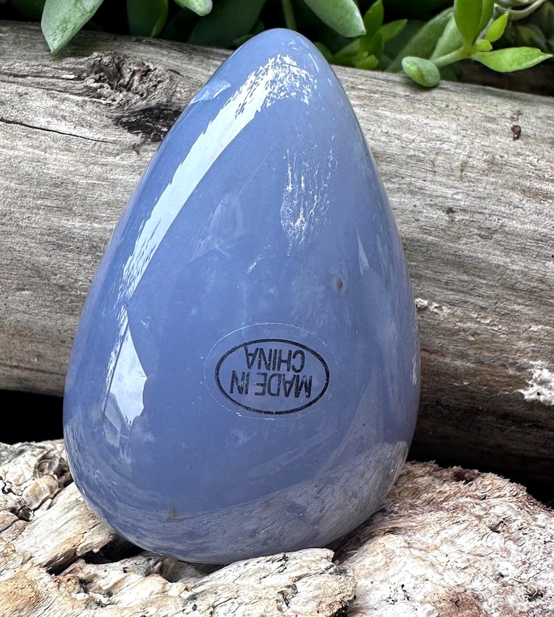 May include: A smooth, teardrop-shaped, pale blue stone. The stone has a glossy finish and a sticker that reads "MADE IN CHINA" in a black circular frame. The stone is resting on a piece of weathered wood.