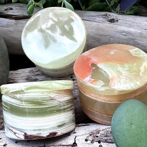 Peut inclure: Trois boîtes décoratives en onyx dans les tons de vert, orange et blanc. Les boîtes sont cylindriques et rectangulaires, avec des couvercles. L'onyx présente des veines naturelles et une finition polie. Les boîtes sont exposées sur une surface en bois.