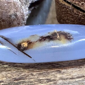 May include: A smooth, oval-shaped blue chalcedony stone featuring a translucent, light periwinkle color. The surface displays a distinct, dark brown dendritic inclusion that stretches across the center, creating a natural, contrasting pattern.