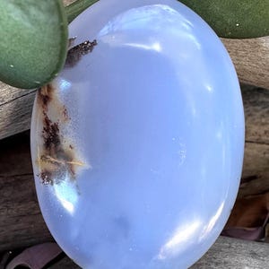 May include: An oval-shaped, smooth, polished gemstone with a translucent light blue color. The stone features a distinct, dark brown, dendritic-like inclusion on the left side, creating a natural contrast against the soft blue surface.