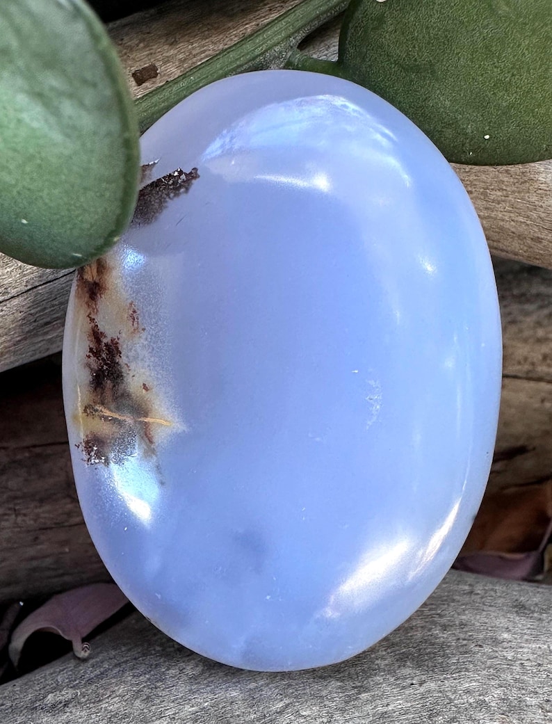 May include: An oval-shaped, smooth, polished gemstone with a translucent light blue color. The stone features a distinct, dark brown, dendritic-like inclusion on the left side, creating a natural contrast against the soft blue surface.
