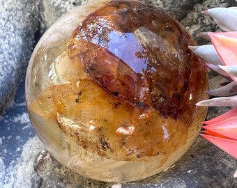May include: A spherical, clear quartz crystal ball containing internal inclusions of golden-yellow and deep reddish-brown mineral deposits. The polished surface of the sphere reflects light, highlighting the natural, irregular textures trapped within the stone.