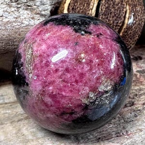 May include: A polished sphere of rhodochrosite, showcasing a vibrant pink hue with black and silver-colored inclusions. The gemstone is smooth and round, reflecting light. The sphere is a decorative item.