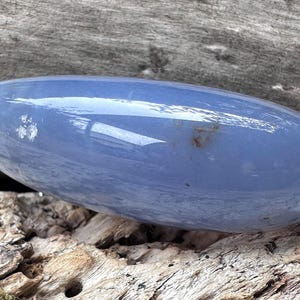 May include: A smooth, oval-shaped blue crystal stone. The polished surface reflects light, and the stone rests on a textured wooden surface. The stone's color is a soft, muted blue, with subtle variations in tone.