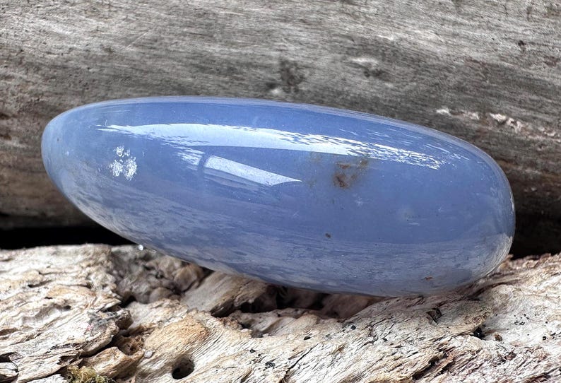 May include: A smooth, oval-shaped blue crystal stone. The polished surface reflects light, and the stone rests on a textured wooden surface. The stone's color is a soft, muted blue, with subtle variations in tone.