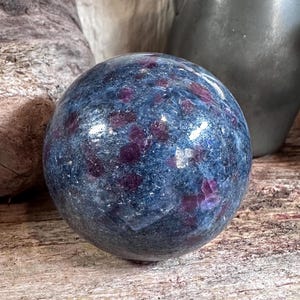 May include: A polished, spherical gemstone with a deep blue base color and scattered patches of dark red. The stone has a smooth, reflective surface and appears to be a decorative item or collectible. The background includes wooden and stone elements.