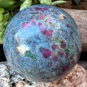 May include: A polished gemstone sphere with a blue-green base color and vibrant magenta inclusions. The sphere is smooth and round, showcasing a natural, earthy aesthetic. The gemstone's surface has a subtle sheen, highlighting its unique patterns and textures.