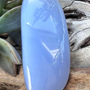 May include: A smooth, oval-shaped, pale blue gemstone. The polished stone reflects light, with subtle variations in color. The background features wood and green foliage, enhancing the stone's natural beauty. This item is ideal for jewelry making.