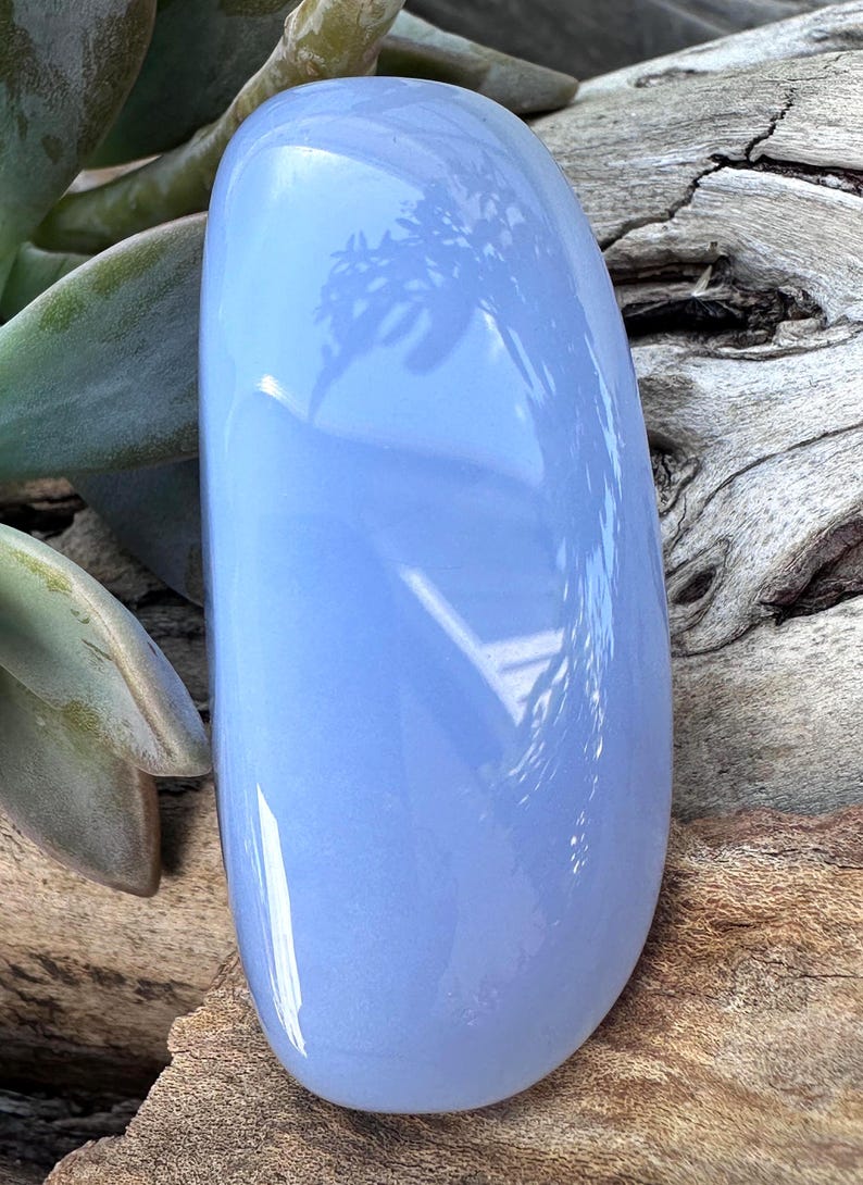 May include: A smooth, oval-shaped, pale blue gemstone. The polished stone reflects light, with subtle variations in color. The background features wood and green foliage, enhancing the stone's natural beauty. This item is ideal for jewelry making.