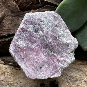 May include: A large, rough, pink ruby crystal with a natural, raw finish. The crystal has a light pink color with a slight purple hue and is covered in small, white inclusions. The crystal is sitting on a piece of wood.