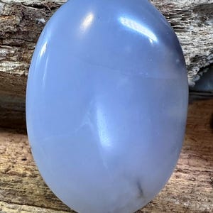 May include: A smooth, oval-shaped blue chalcedony gemstone with a polished, translucent surface. The stone displays a soft, light blue hue with subtle internal cloud-like inclusions and a glossy finish that reflects light across its rounded top.