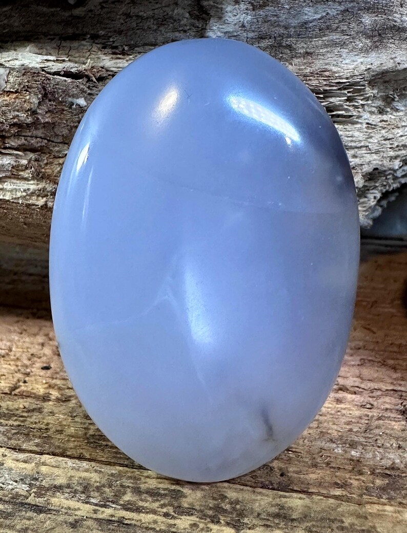 May include: A smooth, oval-shaped blue chalcedony gemstone with a polished, translucent surface. The stone displays a soft, light blue hue with subtle internal cloud-like inclusions and a glossy finish that reflects light across its rounded top.