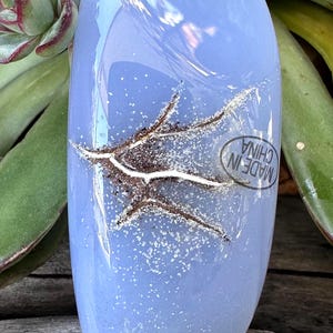 May include: A light blue glass decorative object with a brown and white design. The object has a smooth, glossy surface and a sticker that says "MADE IN CHINA". The object is surrounded by green plants.