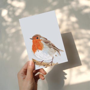 Op de afbeelding: Een aquarel schilderij van een roodborstje op een tak. De vogel heeft een feloranje borst, een bruine rug en een witte buik. Het kunstwerk staat op een rechthoekige kaart met een lichtgrijze achtergrond.