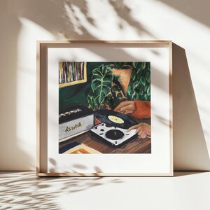 May include: A framed art print depicting a scene with a white record player, a black vinyl record, and a black amplifier with the text Marshall. The composition includes green potted plants, a wooden surface, and a person wearing an orange sweater.
