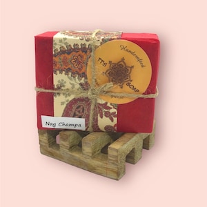 May include: A red and white bar of soap with a brown label that says "Handcrafted Soap" and "Nag Champa". The soap is wrapped in twine and is sitting on a wooden soap dish.
