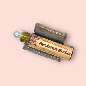 May include: A brown glass roller bottle with a silver roller ball. The label is yellow and brown with the text "Patchouli Amber" and "Ingredients: grape seed oil, patchouli oil, fragrance." The bottle is sitting on a ceramic holder.