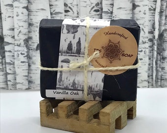 Op de afbeelding: Een zwarte zeep, met het label "Vanilla Oak", is verpakt in papier met patronen en vastgebonden met touw. Een rond, bruin label leest "Handcrafted TTS SOAP". De zeep ligt op een kleine houten zeepbakje.