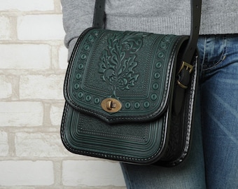 May include: A dark green leather crossbody bag with an intricate embossed floral design. The bag has a black strap and a brass buckle.