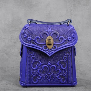 Könnte beinhalten: Eine kleine, violette Ledertasche mit aufwendigen, geprägten Blumenmustern und einem Messingverschluss.
