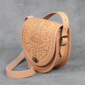May include: A tan leather crossbody bag with a floral embossed design. The bag features a long adjustable strap, a top handle, and a metal clasp closure. The bag's design includes a detailed floral pattern and decorative border.