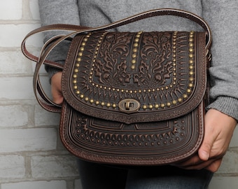 May include: A brown leather crossbody bag with intricate embossed floral designs and gold-toned studs. The bag has a flap closure with a metal buckle.