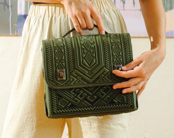 May include: A woman wearing a black vest top and beige trousers is holding a dark green satchel bag with an intricate geometric pattern.
