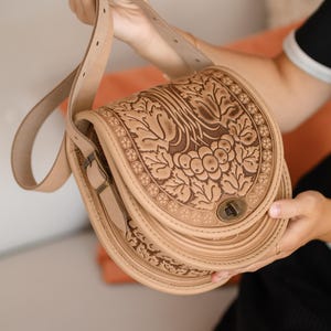 May include: A tan leather saddlebag with a floral design. The bag features a long adjustable strap and a metal clasp closure. The front of the bag is decorated with a detailed embossed pattern of leaves and grapes.