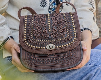 May include: A brown leather satchel with intricate floral tooling and gold-toned studs. The bag has a flap closure with a metal buckle and a shoulder strap.