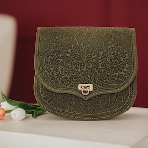 May include: Olive green crossbody bag with a floral embossed design. The bag features a curved flap with a metal clasp closure. The bag is sitting on a white surface with a red background. A few flowers are in the foreground.