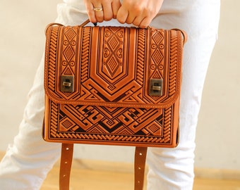 May include: A brown leather satchel with intricate geometric patterns and a strap. The bag is being held by a person wearing white trousers and a striped shirt.